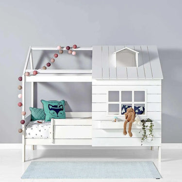 White wooden house-themed children's bed with a small roof, window, and railing. Decorated with fairy lights, cushions, and a plush bunny.
