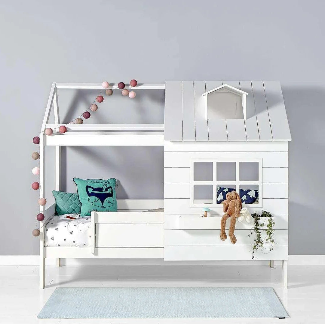 White wooden house-themed children's bed with a small roof, window, and railing. Decorated with fairy lights, cushions, and a plush bunny.
