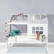 White wooden house-themed children's bed with a small roof, window, and railing. Decorated with fairy lights, cushions, and a plush bunny.