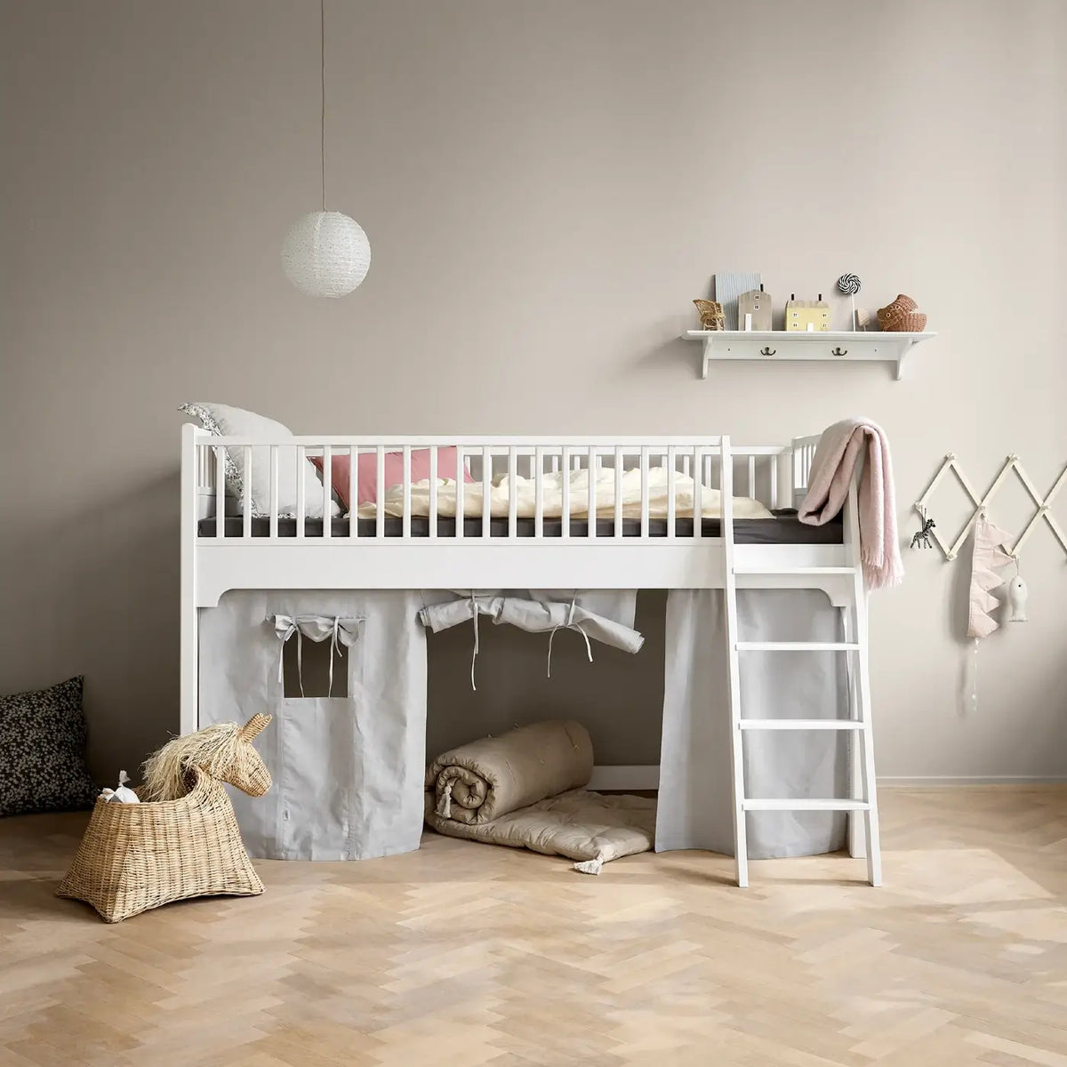 A white wooden loft bed with a ladder, dressed in neutral bedding, set in a child's bedroom with a beige wall, light wood herringbone floor, and a gray fabric play tent underneath.